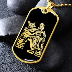 May include: A gold-colored dog tag pendant with a black center featuring a detailed gold illustration of a seated figure holding a bird. The pendant hangs from a gold ball chain, resting on dark gray stones.