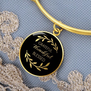 May include: Gold-colored bangle bracelet with a round black pendant. The pendant has the words "Grateful, Thankful, Blessed" in gold script, framed by gold leaf designs. The bracelet is set against a light blue and gold lace background.