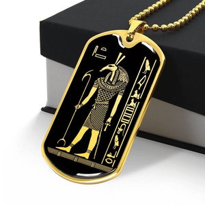 Puede incluir: Un collar con una placa de identificación dorada y un fondo de esmalte negro. El colgante presenta una imagen detallada del dios egipcio Anubis, rodeado de jeroglíficos.