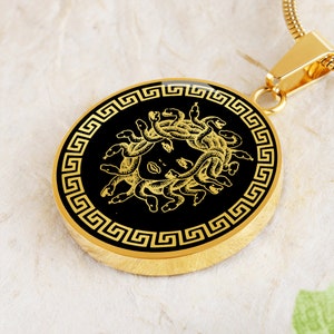May include: Gold pendant necklace with a black circular charm featuring a gold image of Medusa, a Greek mythological figure with snakes for hair, surrounded by a gold Greek key border.