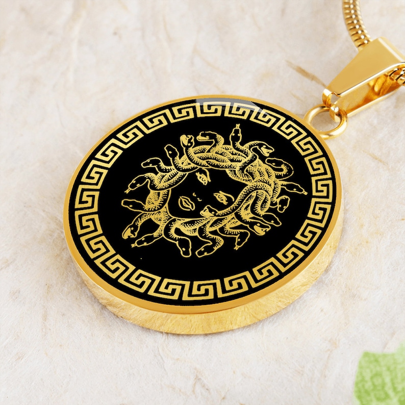 May include: Gold pendant necklace with a black circular charm featuring a gold image of Medusa, a Greek mythological figure with snakes for hair, surrounded by a gold Greek key border.