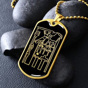 Op de afbeelding: Een goudkleurige dogtag-hanger met een zwart midden en een gegraveerd Egyptisch hiërogliefenontwerp. De hanger hangt aan een goudkleurige kogelketting. De tag is rechthoekig met afgeronde hoeken.