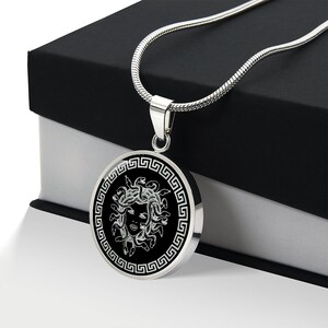 May include: A silver chain necklace with a round pendant featuring a black and white image of Medusa's head surrounded by a Greek key border.