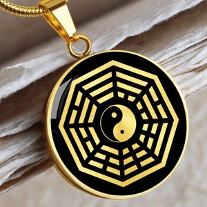 Puede incluir: Un collar dorado con un colgante redondo que presenta un s&iacute;mbolo yin yang negro dentro de un dise&ntilde;o de ocho trigramas, o Bagua, dorado.