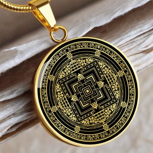 May include: Gold pendant necklace with a black and gold mandala design. The mandala features a square with a complex geometric pattern inside. The pendant is surrounded by a circle of text.