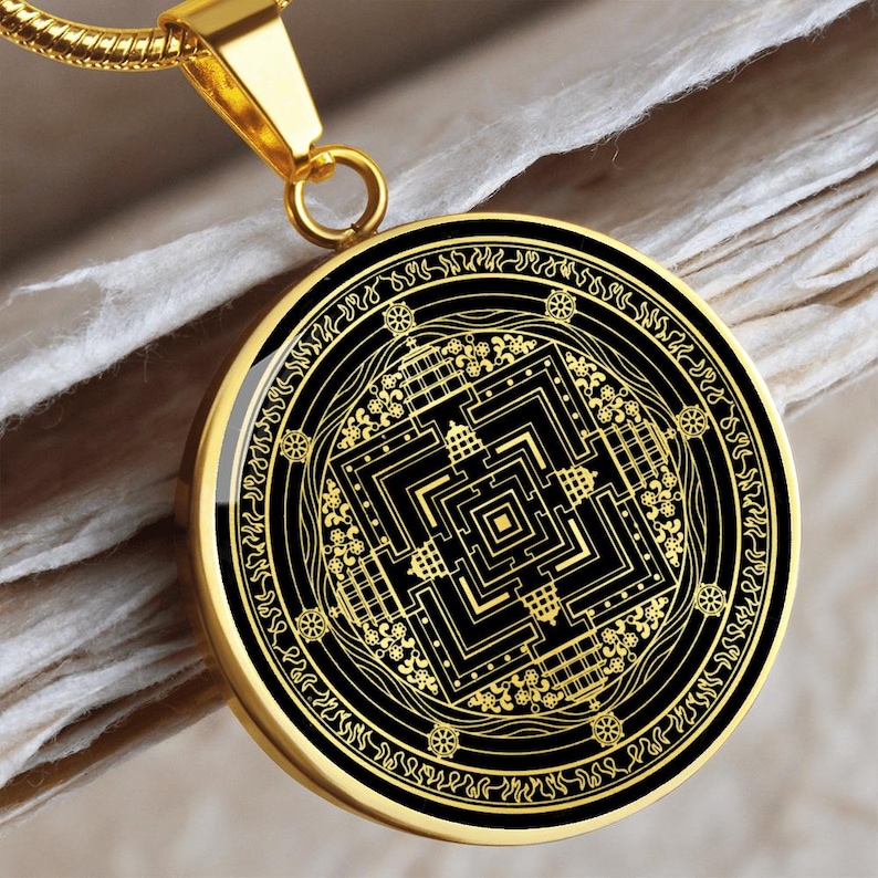 May include: Gold pendant necklace with a black and gold mandala design. The mandala features a square with a complex geometric pattern inside. The pendant is surrounded by a circle of text.