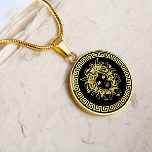 May include: A gold necklace with a round pendant featuring a black and gold image of Medusa, the Greek mythological figure with snakes for hair, surrounded by a Greek key border.