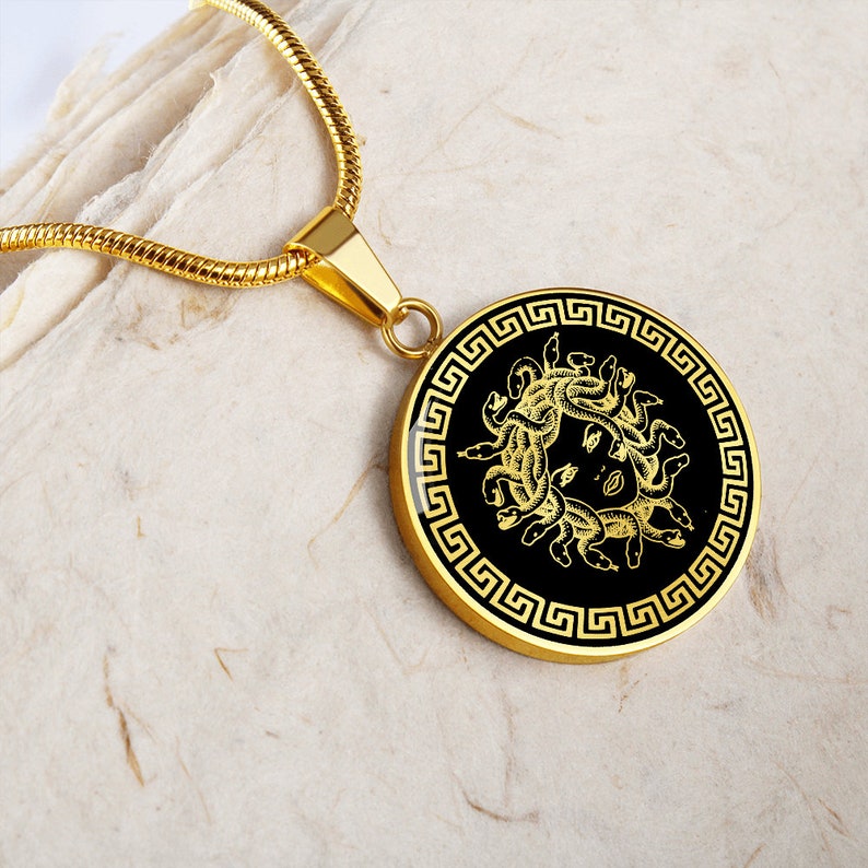May include: A gold necklace with a round pendant featuring a black and gold image of Medusa, the Greek mythological figure with snakes for hair, surrounded by a Greek key border.