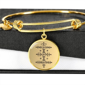 May include: A gold bangle bracelet with a round charm featuring a black and white symbol. The charm is attached to the bracelet with a gold chain.