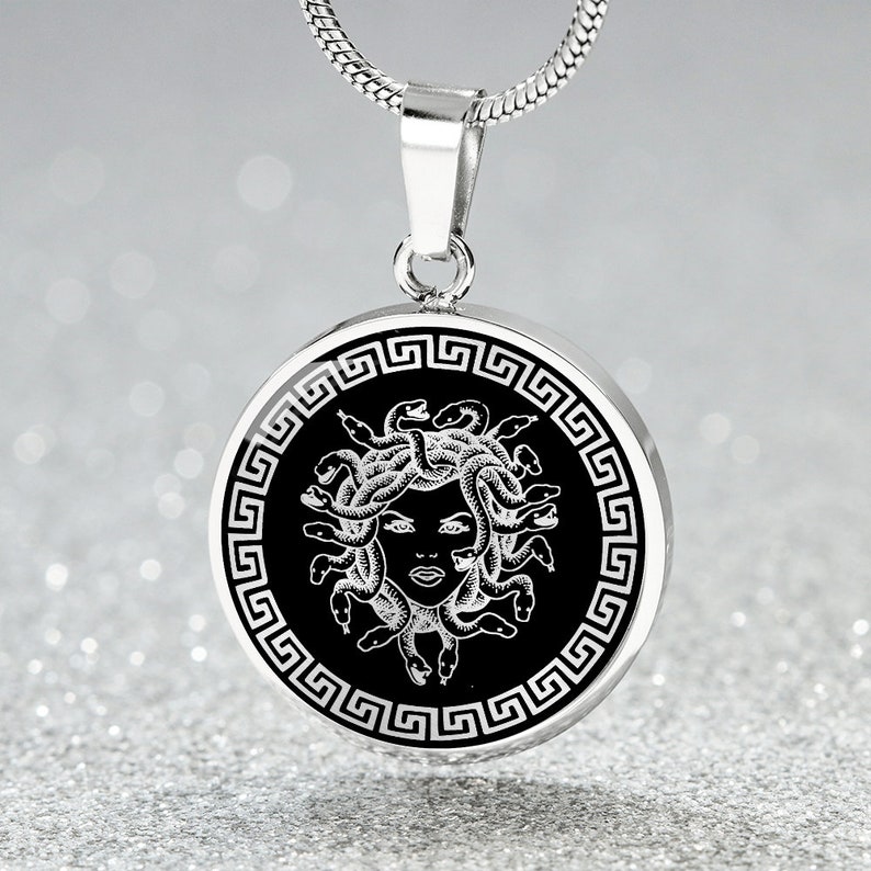 May include: A silver necklace with a round pendant featuring a black and white image of Medusa, the Greek mythological figure with snakes for hair, surrounded by a Greek key border.