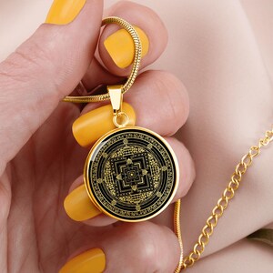 May include: Gold necklace with a round pendant featuring a black and gold geometric design. The pendant is a mandala with a square in the center and intricate details.