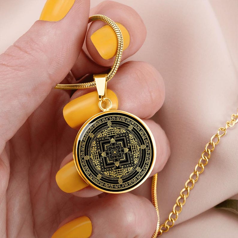 May include: Gold necklace with a round pendant featuring a black and gold geometric design. The pendant is a mandala with a square in the center and intricate details.