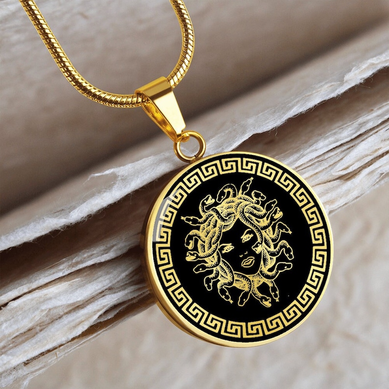 May include: A gold chain necklace with a round pendant featuring a black and gold image of Medusa, the Greek mythological figure with snakes for hair, surrounded by a Greek key border.