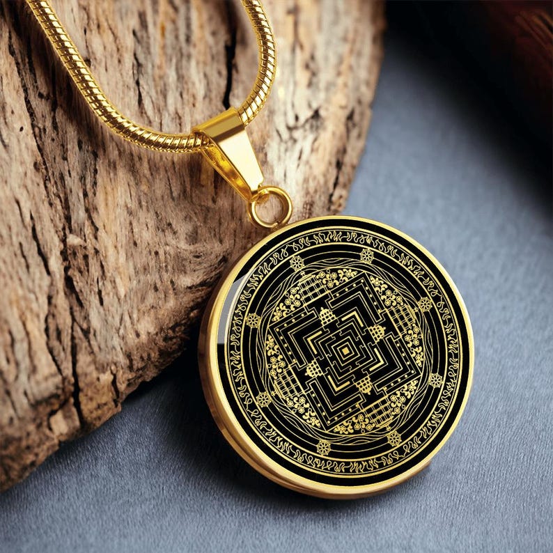 May include: A gold chain necklace with a round pendant featuring a black and gold mandala design. The mandala is intricate and detailed, with a square shape in the center and a circular border.