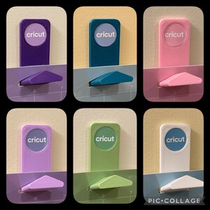 May include: Six Cricut cutting tools in different colors: purple, teal, pink, lavender, green, and white. Each tool has a small, rectangular base with the word "cricut" printed on it. The tools are arranged in two rows of three, with the base of each tool facing the viewer.