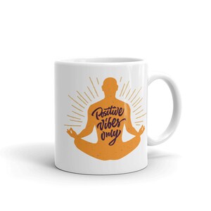 Puede incluir: Taza de caf&eacute; de cer&aacute;mica blanca con una silueta de una persona en una postura de yoga. La persona es naranja y el texto "Positive Vibes Only" est&aacute; escrito en morado dentro de la silueta.