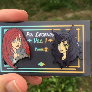May include: Two enamel pins featuring cartoon characters. One pin shows a woman with red hair and a skull on her belt. The other pin depicts a woman with black hair and cat ears. The pins are on a black card with the text "Pin Legends Vol. 1".
