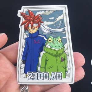 Peut inclure: Un autocollant représentant deux personnages de dessin animé, une personne aux cheveux roux portant une veste bleue et une grenouille verte portant une veste verte. L'autocollant a une bordure blanche et le texte "2300 AD" en bas.