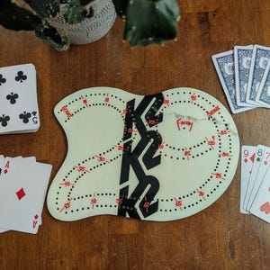 May include: A cribbage board with a black and white design, surrounded by playing cards. The board is off-white with red markings and the word "Finish" in red. A deck of cards is visible, with the five of clubs on top.