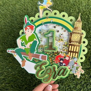 Puede incluir: Un adorno para pastel de Peter Pan de color verde y dorado con una figura de Peter Pan volando en un gancho, Campanita, una torre del reloj Big Ben y el nombre "Ezra" en purpurina dorada. El adorno está decorado con purpurina verde y dorada.