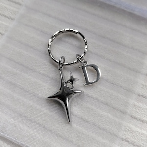 May include: Silver-toned keychain featuring a starburst charm and a letter "D" pendant. The keychain has a textured ring and is likely made of metal. The starburst charm has a four-pointed design.