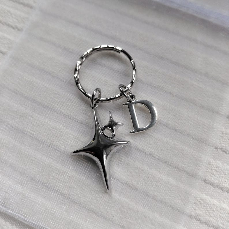 May include: Silver-toned keychain featuring a starburst charm and a letter "D" pendant. The keychain has a textured ring and is likely made of metal. The starburst charm has a four-pointed design.
