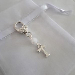 May include: Silver charm with a heart-shaped clasp, a white bead, and a silver letter T. The charm is hanging from a white organza bag.