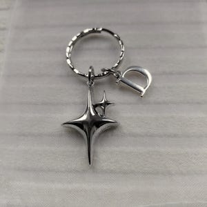 May include: Silver-toned keychain featuring a starburst charm and a letter "D" pendant. The keychain has a textured ring and a small star charm. The design is sleek and modern, suitable for everyday use.
