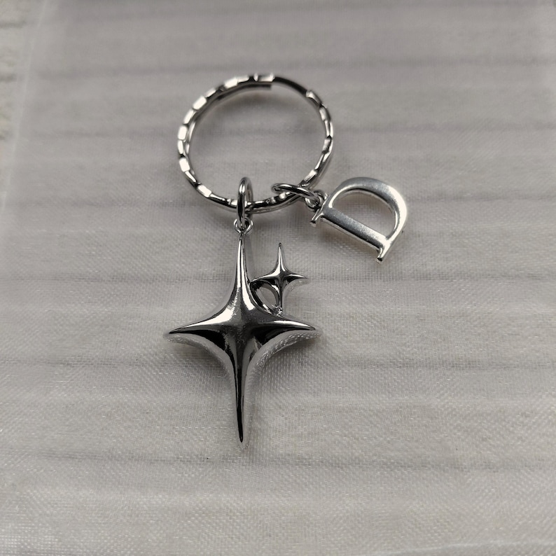 May include: Silver-toned keychain featuring a starburst charm and a letter "D" pendant. The keychain has a textured ring and a small star charm. The design is sleek and modern, suitable for everyday use.