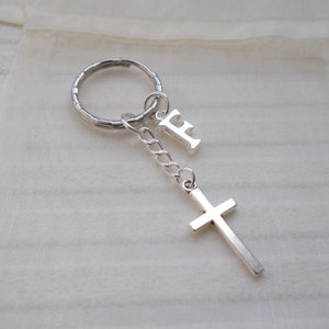 May include: A silver-toned metal keychain featuring a circular split ring, a short chain, a capital letter F charm, and a simple cross pendant. The components are connected by small jump rings, creating a dangling design with a metallic finish.