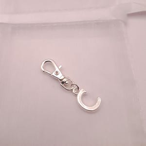 May include: A silver-colored lobster clasp with a swivel base and a small, curved charm. The clasp is designed to attach to a key ring or other accessories. The background is a soft, off-white color.