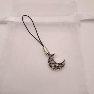May include: A silver-coloured crescent moon charm with a decorative filigree design and stars. The charm is attached to a black cord with a silver-coloured metal clasp. The background is a soft white fabric.