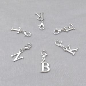 May include: Silver alphabet charms with lobster claw clasps. The letters T, R, E, K, N, and B are shown. These charms are suitable for jewellery making or personalising accessories.