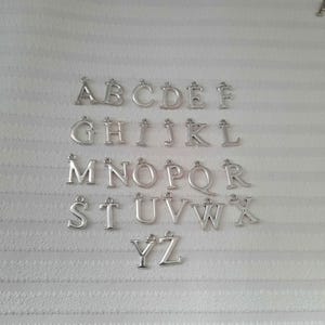 May include: Silver alphabet charms, capital letters A through Z, arranged on a white, textured surface. Each letter has a small loop at the top for attaching to jewelry or other crafts. The letters are evenly spaced.