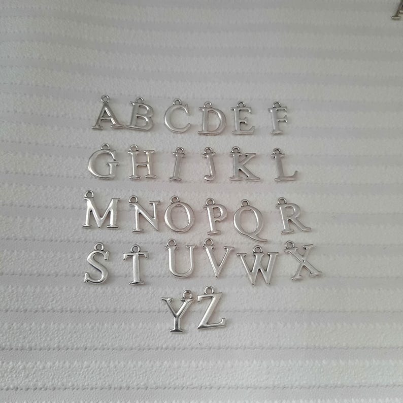 May include: Silver alphabet charms, capital letters A through Z, arranged on a white, textured surface. Each letter has a small loop at the top for attaching to jewelry or other crafts. The letters are evenly spaced.