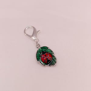 May include: A silver charm depicting a red and black ladybug on a green leaf. The ladybug has black spots and is set against a detailed green leaf. The charm is attached to a silver clasp, suitable for a bracelet or necklace.