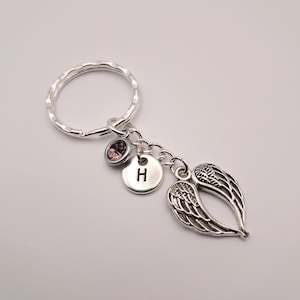 May include: Silver-toned keychain featuring a round key ring, a small pink gemstone, a circular charm with the letter "H", and a pair of detailed angel wings. The keychain is designed with a delicate and elegant aesthetic.