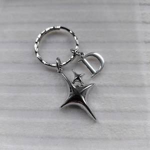 May include: Silver-toned keychain featuring a stylized starburst design and a letter "D" charm. The keychain includes a split ring for attaching keys and a small star accent. The metal has a polished finish.