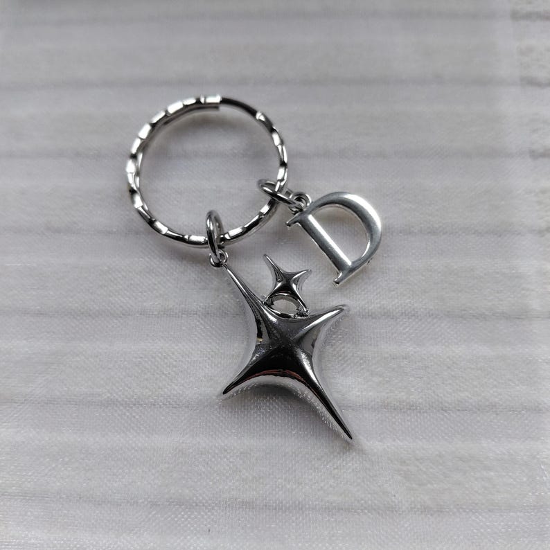 May include: Silver-toned keychain featuring a stylized starburst design and a letter "D" charm. The keychain includes a split ring for attaching keys and a small star accent. The metal has a polished finish.