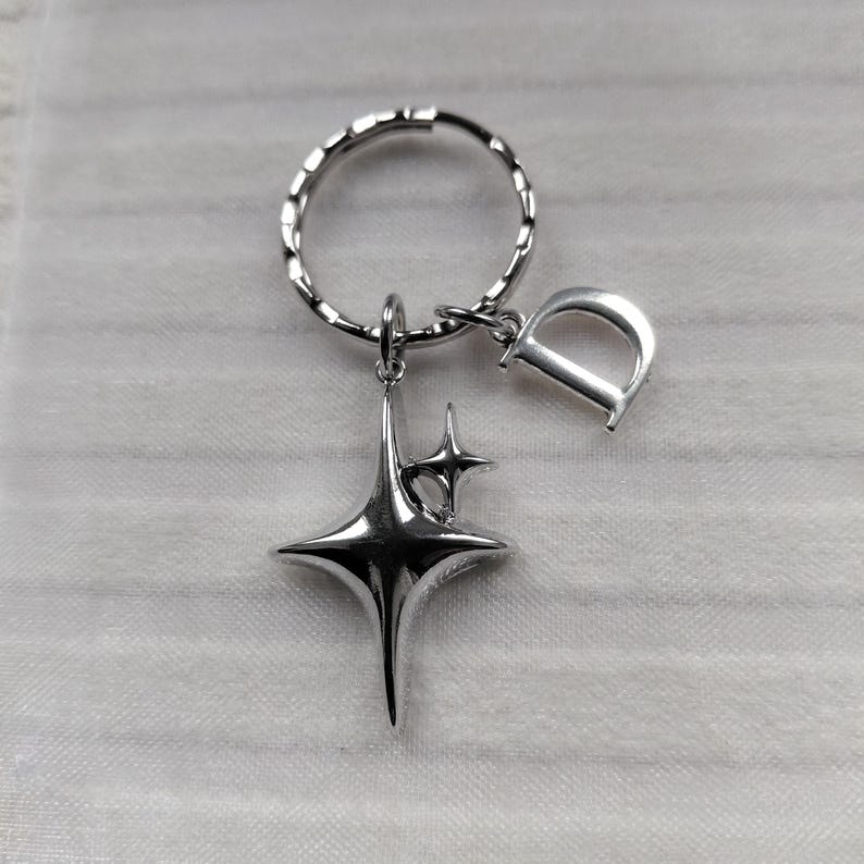May include: Silver-toned keychain featuring a starburst charm and a letter "D" charm. The starburst has a three-dimensional design with multiple points. The keychain ring is a twisted design.