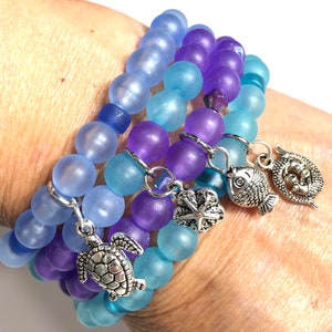 Könnte beinhalten: Drei Armbänder mit blauen und violetten Perlen. Die Armbänder haben runde, mattierte Perlen und silberne Anhänger, darunter eine Schildkröte, ein Seestern, ein Fisch und ein dekoratives Oval. Die Armbänder sind so konzipiert, dass sie zusammen getragen werden können.