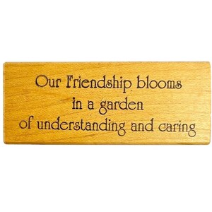 以下が含まれることがあります： 「Our friendship blooms in a garden of understanding and caring」と刻まれた長方形の木製プラーク。 木材は明るい茶色で、木目がはっきりと見えます。