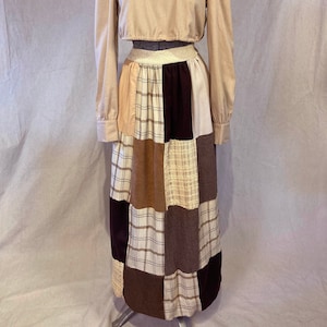 May include: A two-piece outfit on a mannequin. The top is a light tan long-sleeved crop top with a scoop neck and a cream-colored scarf. The skirt is a patchwork design with squares of various brown and tan fabrics.