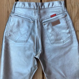 Peut inclure: Jean Wrangler m&eacute;tallis&eacute; argent&eacute;. Le jean a deux poches arri&egrave;re avec les coutures du logo Wrangler, un patch en cuir marron et une &eacute;tiquette Wrangler rouge. Le jean est fait d'un mat&eacute;riau brillant et r&eacute;fl&eacute;chissant.