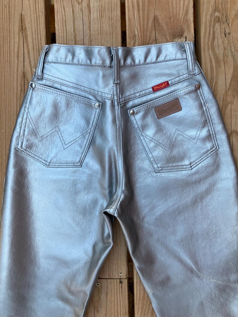 Peut inclure: Jean Wrangler m&eacute;tallis&eacute; argent&eacute;. Le jean a deux poches arri&egrave;re avec les coutures du logo Wrangler, un patch en cuir marron et une &eacute;tiquette Wrangler rouge. Le jean est fait d'un mat&eacute;riau brillant et r&eacute;fl&eacute;chissant.