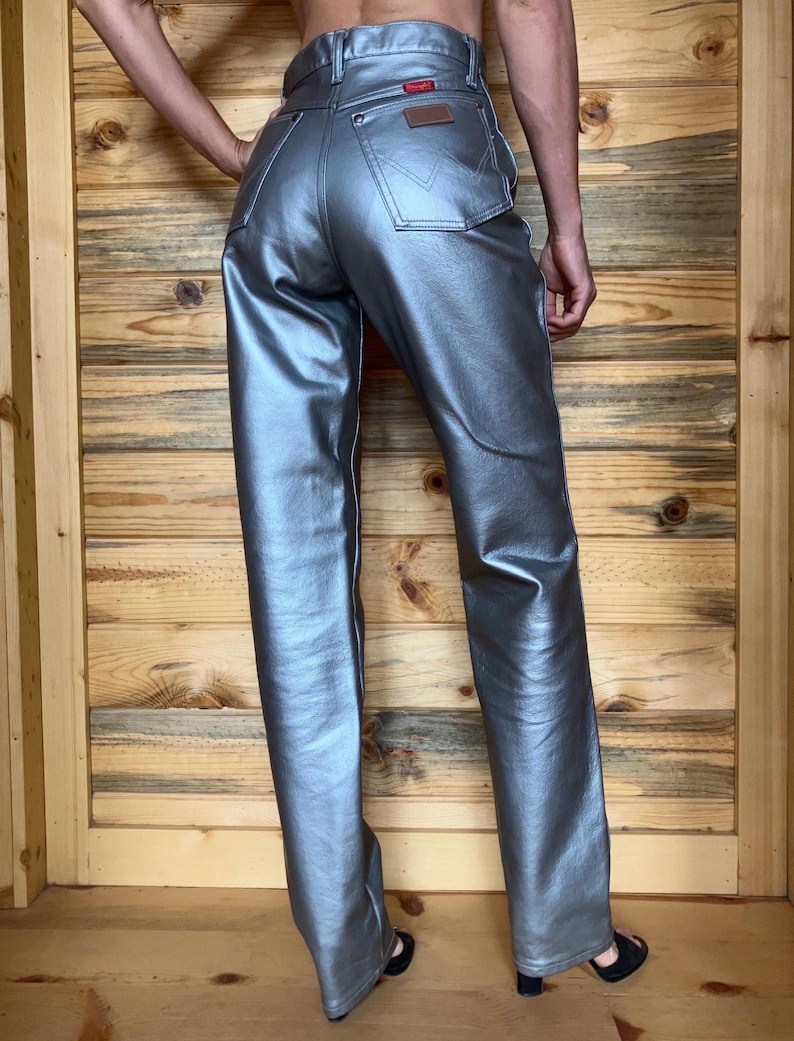 Peut inclure: Un jean Wrangler argent&eacute; m&eacute;tallis&eacute; &agrave; taille haute et cinq poches classiques. Le jean pr&eacute;sente un patch en cuir marron et une &eacute;tiquette Wrangler rouge. Le pantalon a une coupe droite et est fait d'un mat&eacute;riau brillant et r&eacute;fl&eacute;chissant.