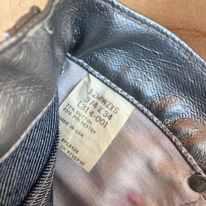 Peut inclure: Gros plan sur un jean en denim m&eacute;tallis&eacute; argent&eacute;. Le jean porte une &eacute;tiquette avec la taille 3/4 X 34 et le texte "MADE IN USA". Le tissu est un m&eacute;lange de coton et de polyester. Le jean est partiellement d&eacute;boutonn&eacute;.