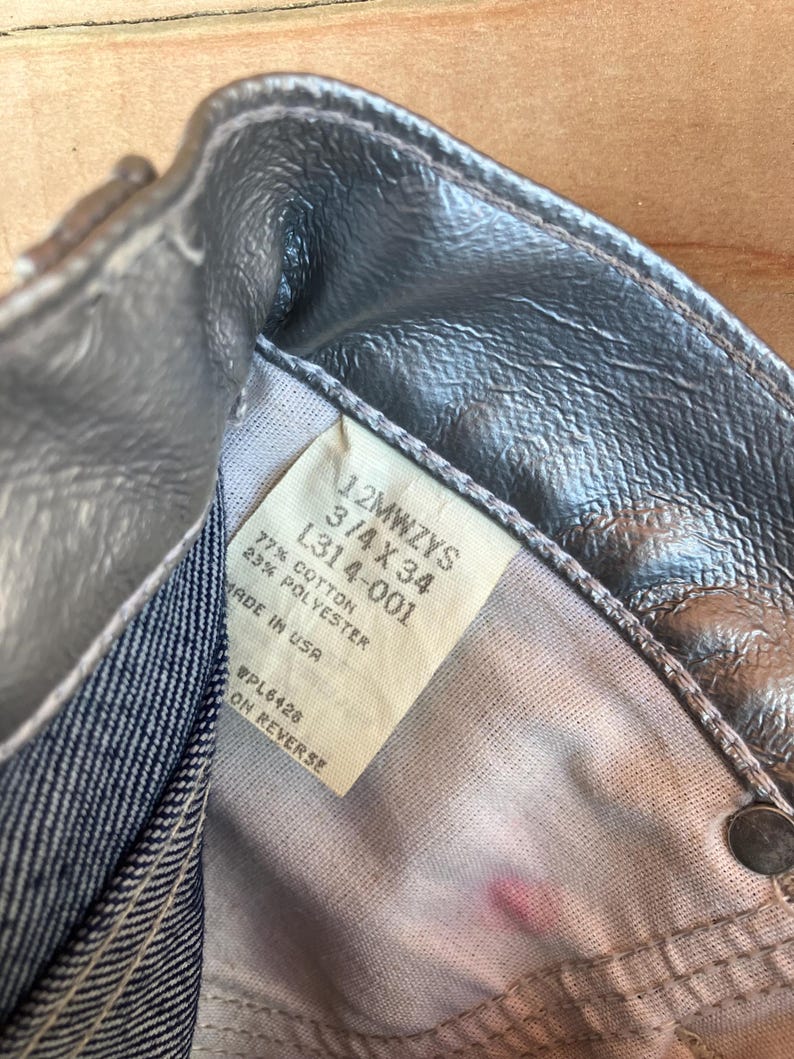 Peut inclure: Gros plan sur un jean en denim m&eacute;tallis&eacute; argent&eacute;. Le jean porte une &eacute;tiquette avec la taille 3/4 X 34 et le texte "MADE IN USA". Le tissu est un m&eacute;lange de coton et de polyester. Le jean est partiellement d&eacute;boutonn&eacute;.