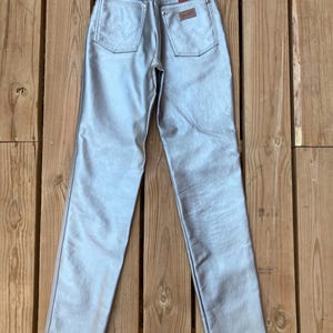 Peut inclure: Jean Wrangler m&eacute;tallis&eacute; argent&eacute; avec une conception classique &agrave; cinq poches. Les poches arri&egrave;re pr&eacute;sentent les coutures Wrangler embl&eacute;matiques. Un patch en cuir marron avec le logo Wrangler est visible sur la ceinture arri&egrave;re. Le jean est pr&eacute;sent&eacute; sur une surface en bois.