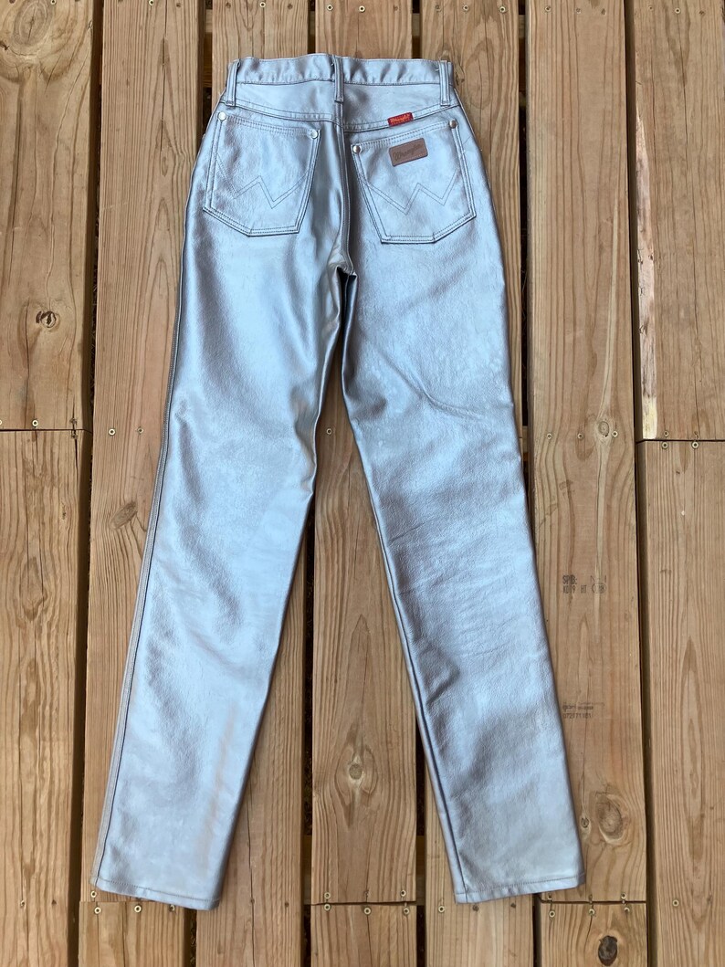 Peut inclure: Jean Wrangler m&eacute;tallis&eacute; argent&eacute; avec une conception classique &agrave; cinq poches. Les poches arri&egrave;re pr&eacute;sentent les coutures Wrangler embl&eacute;matiques. Un patch en cuir marron avec le logo Wrangler est visible sur la ceinture arri&egrave;re. Le jean est pr&eacute;sent&eacute; sur une surface en bois.